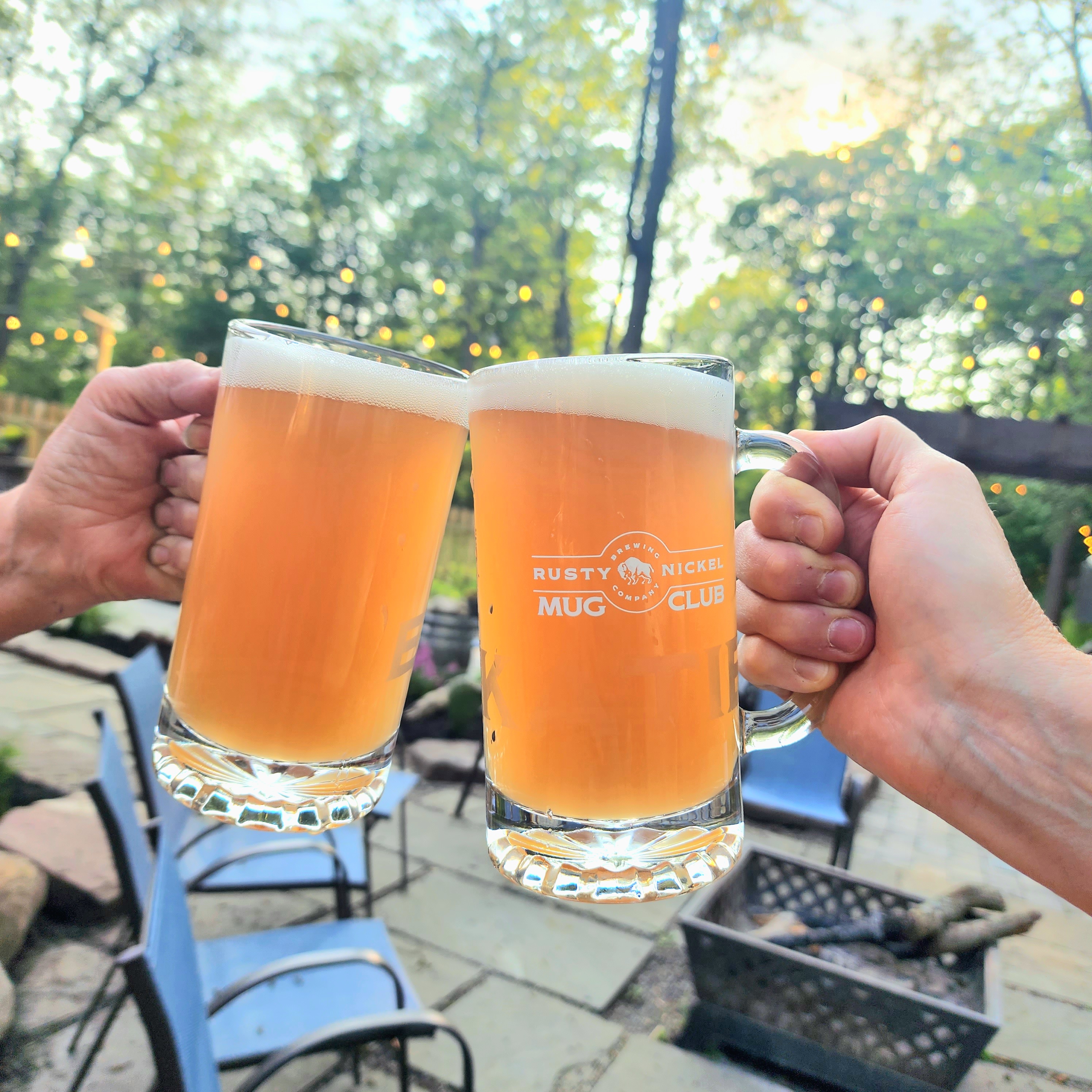Two glass mugs are held up together for a toast on the patio. A summer scene of patio chairs, trees, and a fire pit are in the background.