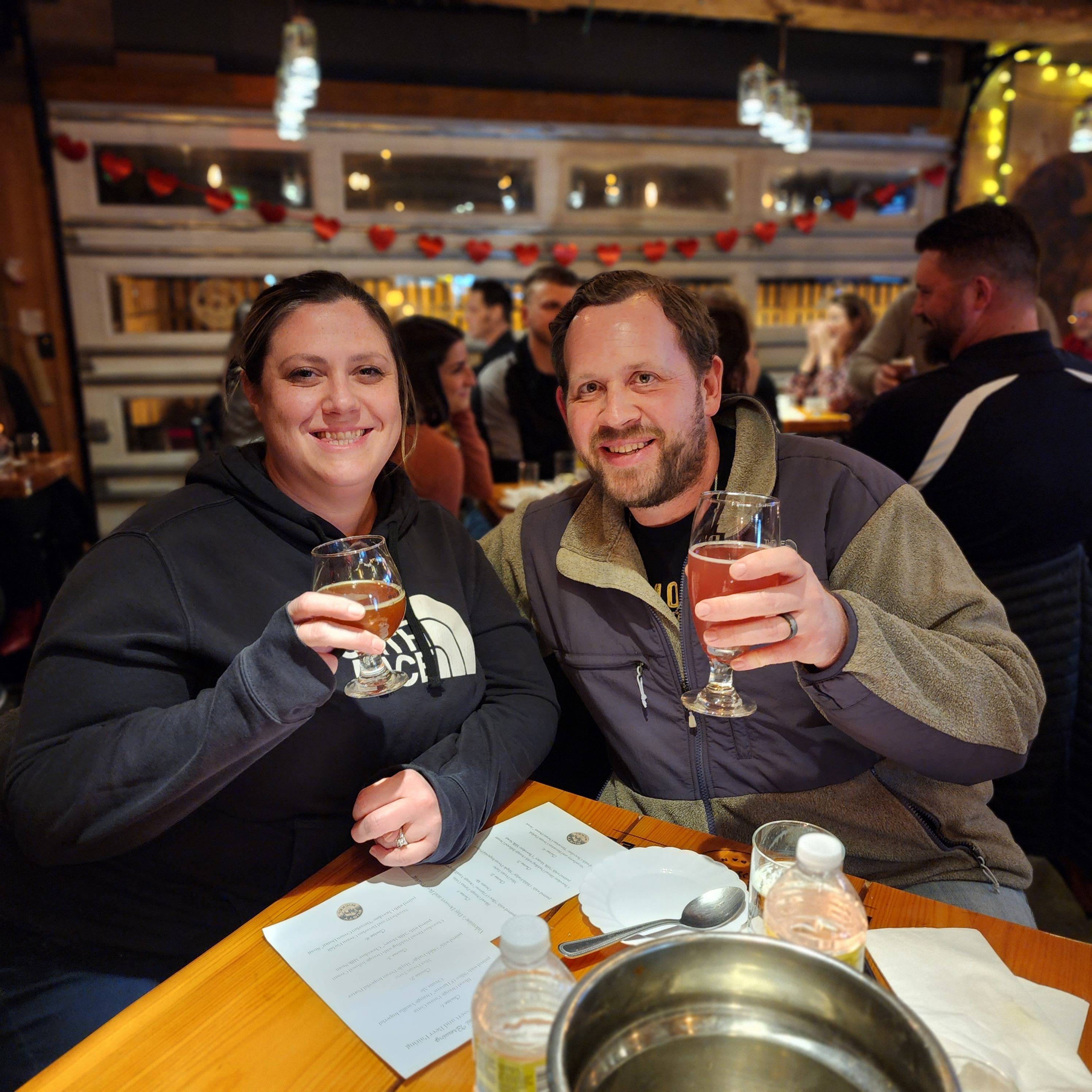 20230214_201826 Woman and Man smile while holding beers at a wooden table. valentines day decorations are behind them