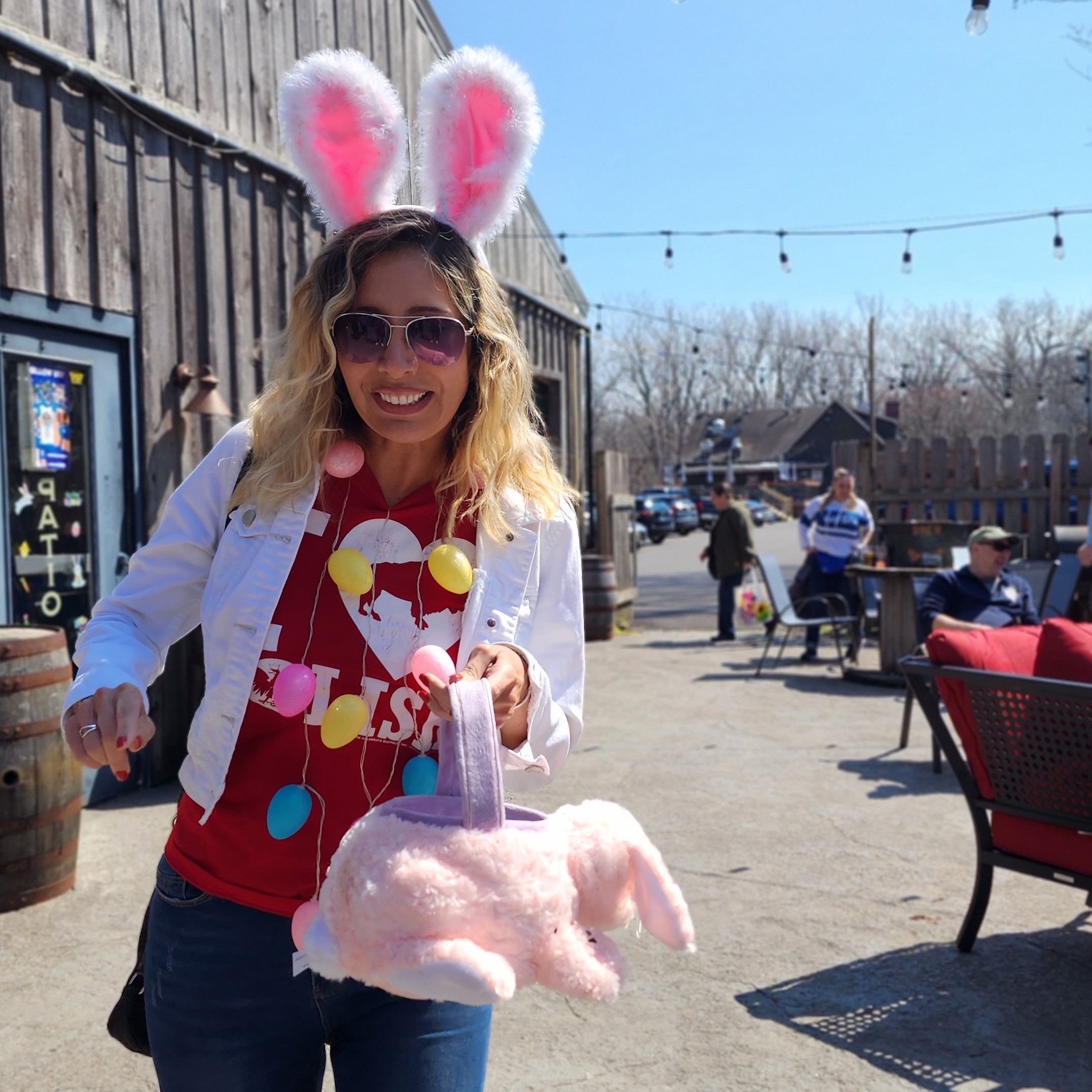 Woman wearing bunny ears holds an easter basket while wearing an easter egg necklace and sunglasses on a patio