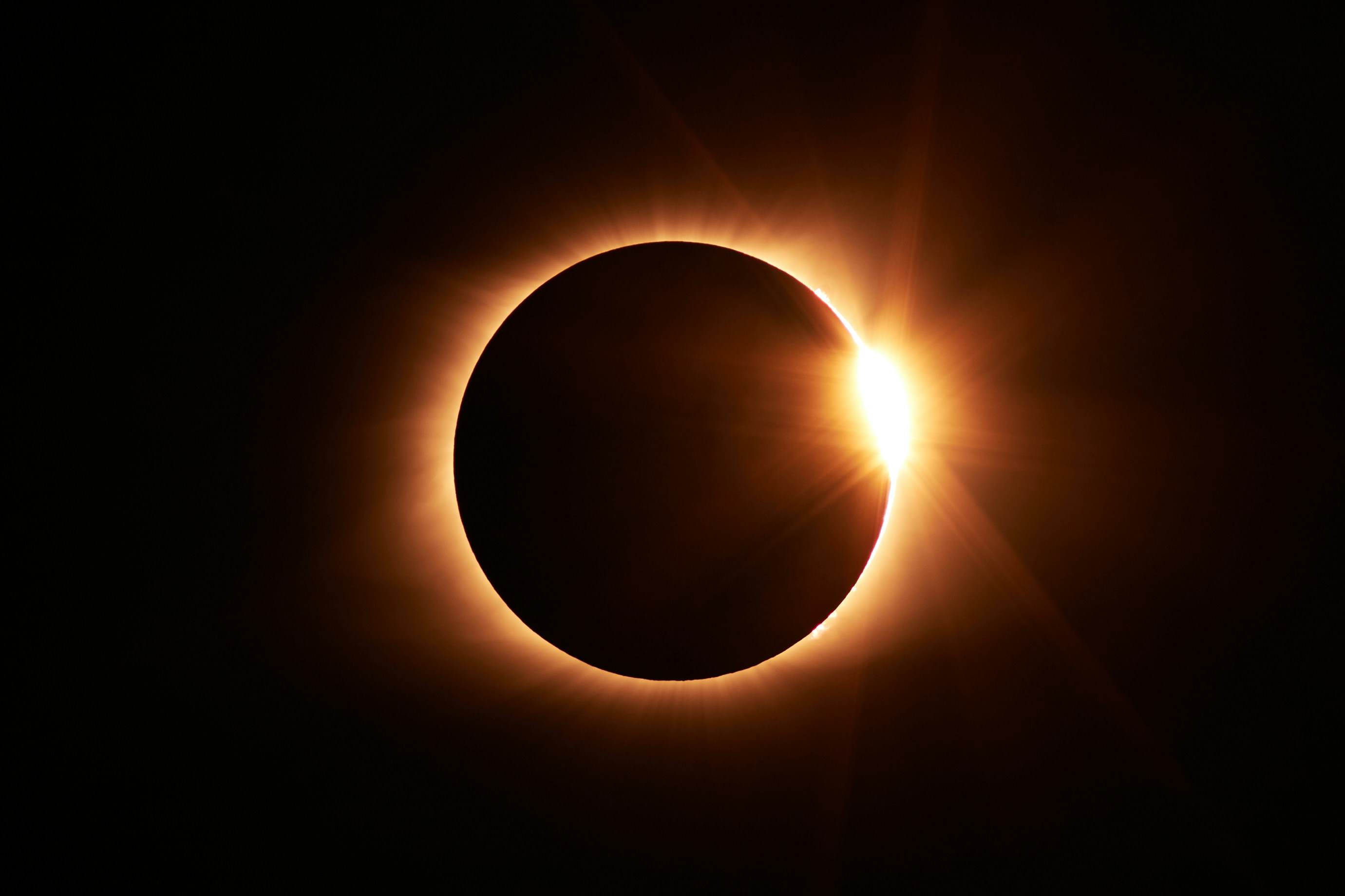 Solar eclipse image shows moon's shadow covering the sun, with bright rays emitting in a ring shape from behind the moon