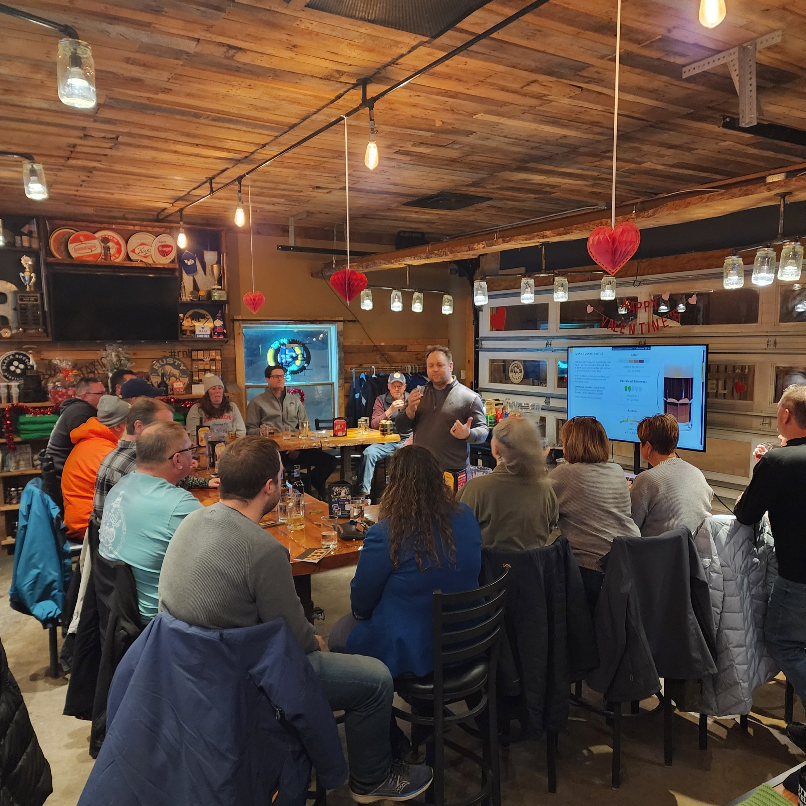 Owner, Jason Havens, instructs a Beer School class. Attendees are seated around tables the rustic Tasting Room. A presentation board is seen in the background.