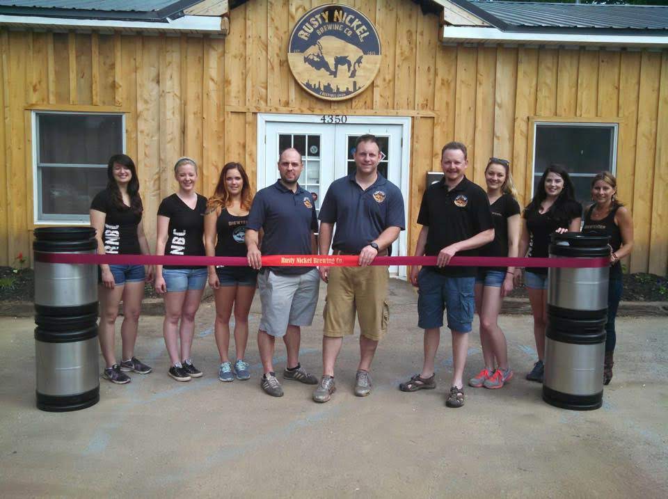 The ribbon cutting ceremony at the Grand Opening of Rusty Nickel Brewing. Owners Jason Havens and David Johnson stand behind the ribbon ready to cut, they are joined by members of their staff in front of the Tasting Room.