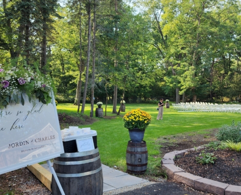a sign welcomes guests to an outdoor wedding ceremony. Chairs are lined up on a lawn surrounded by tall trees and whiskey barrels decorated with flowers.