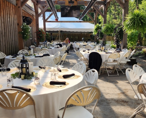 A large, covered pergola stands over several tables set for a wedding reception. The tables are round, with linens, centerpieces, and programs.