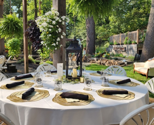 a wedding table setting. White linen on a round table, with a candlelit lantern as the centerpiece. each setting has a gold charger, rolled silverware, and a program. the table is outside. Hanging plants and a garden can be seen in the distance.