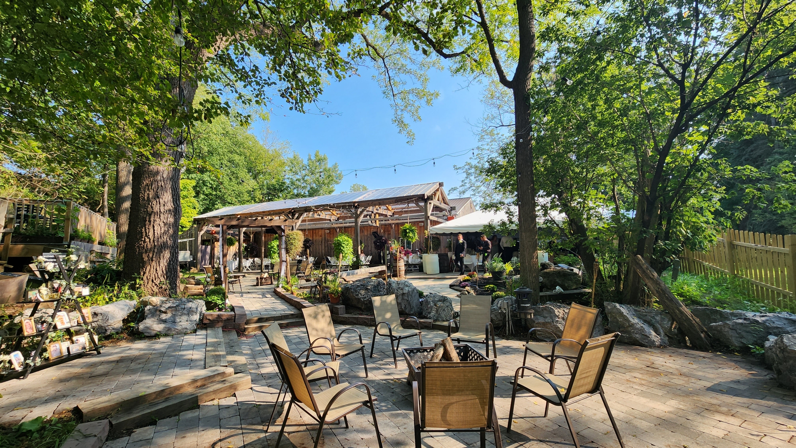 A view of the patio and beer garden at Rusty Nickel Brewing. Patio chairs surround a fire pit in a paved section in the foreground. Garden beds and walkways sprawl the garden area. A large tent and a timber frame pergola are seen in the background adjacent to the building.