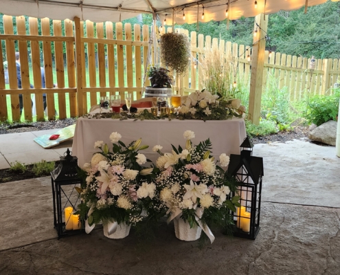 A sweetheart table is set for a new bride and groom. In front of the table, two large lanterns are seen next to several large floral arrangements.