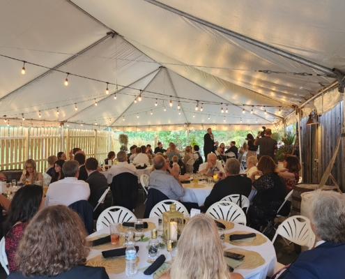 a long white tent covers a wedding reception. guests are seated at their dining tables. A man gives a speech at the far end. Patio lights are strung along the canopy.