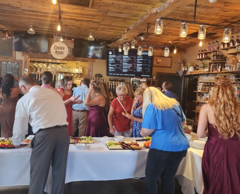 Guests help themselves to appetizers and hors d'ouevres in the Tasting Room while other guests order drinks at the bar behind them. The room is rustic, with lots of exposed wood and warm lighting made of mason jars.