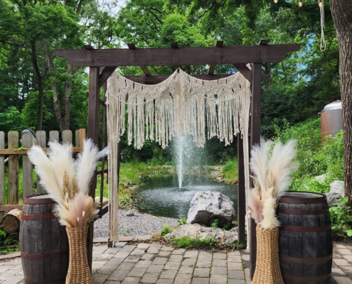 An arbor sits on pavers in front of a pond with a fountain. the wooden structure is decorated with macrame drapery and two large vases of pampas grass are set on either side.