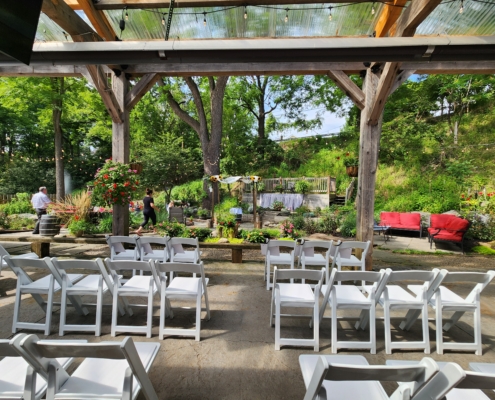 chairs are set in rows for a wedding ceremony. The chairs are under a covered pergola, overlooking a central ceremony in the garden