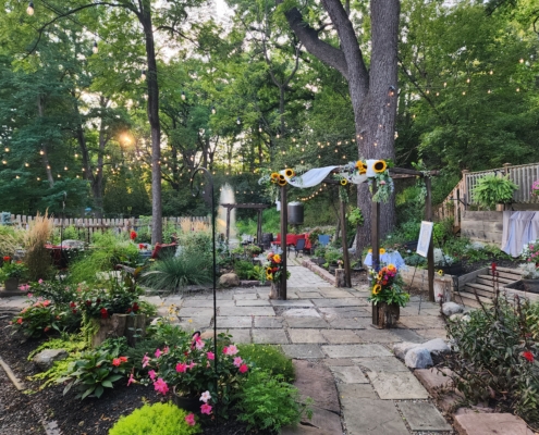 a wooden chuppah is decorated with drapery and sunflowers, and sits on pavers in the garden. The sunsets in the background over a pond with a fountain.