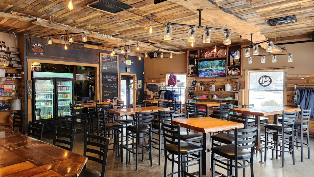 The rustic Tasting Room inside Rusty Nickel Brewing. The room is decorated with live edge wood elements. The room shows several high-top tables and chairs. Merchandise and a beer to go cooler line the walls.
