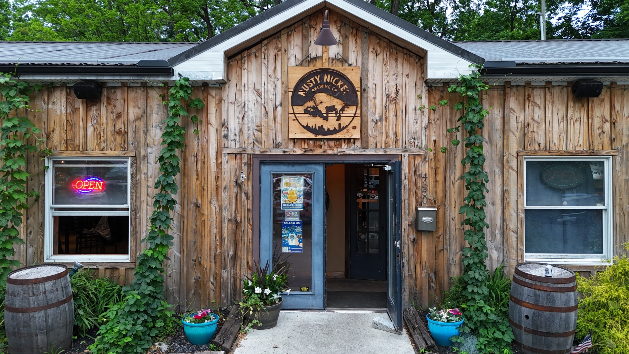 The front entrance to Rusty Nickel Brewing's Tasting Room. A one-story building with wood siding. A set of doors in centered on two sets of windows. A round wooden sign hangs above the front door.