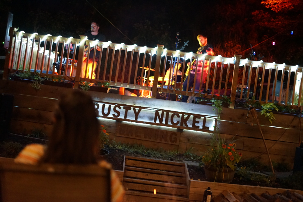 Musicians perform on the stage in the beer garden at night. guests watch while enjoying a fire.