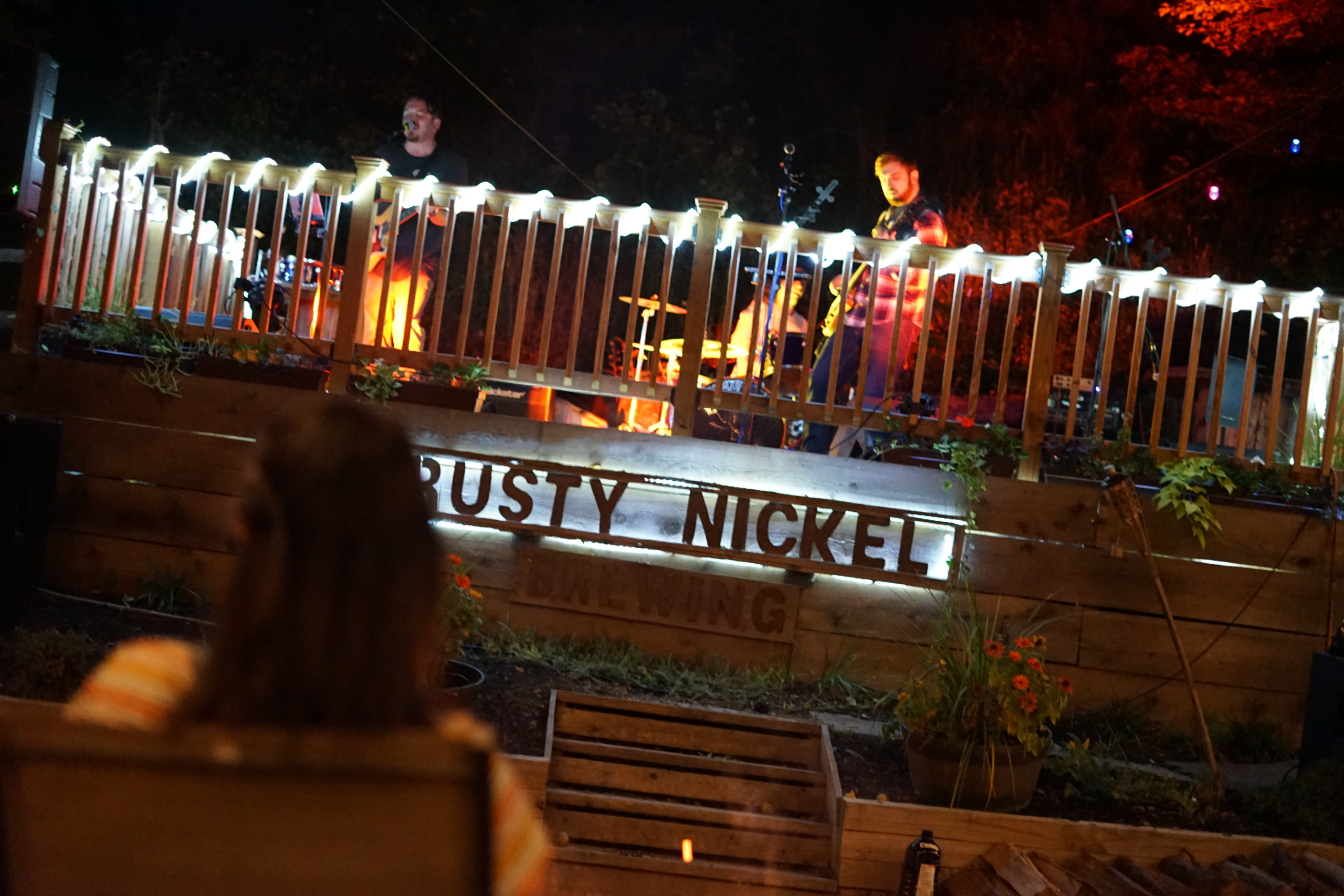 Musicians perform on the stage in the beer garden at night. guests watch while enjoying a fire.