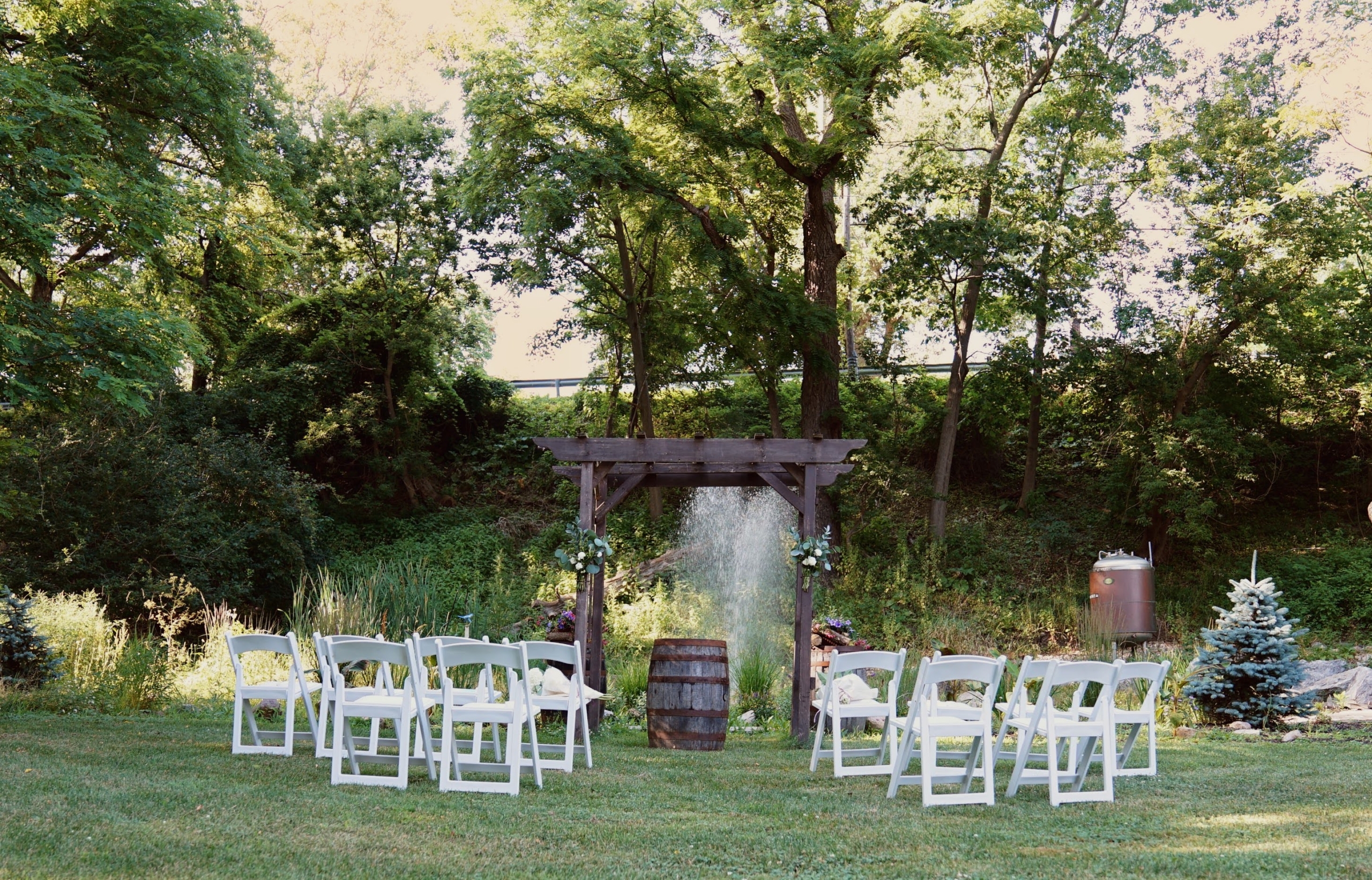 A wooden arbor is set on a lawn in front of a fountain. The background has tall, leafy trees. A whiskey barrel sits under the arbor, and chairs are organized in rows on either side of the aisle.