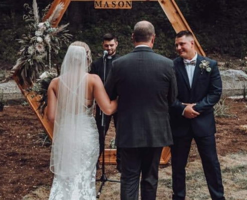 a father walks his daughter, a bride, to the alter, where her groom awaits. a wooden arbor decorated with flowers sits behind them.