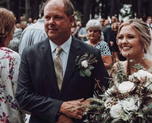 a bride walks down the aisle with a large floral bouquet escorted by her father in a suit.