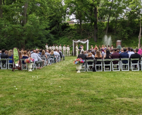 A wedding ceremony takes place on a large lawn, with a background of large trees and shrubbery.