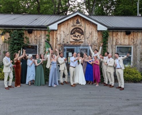 a wedding party cheers as a newlywed couple sprays champagne in celebration. The group is standing in front of Rusty Nickel Brewing.