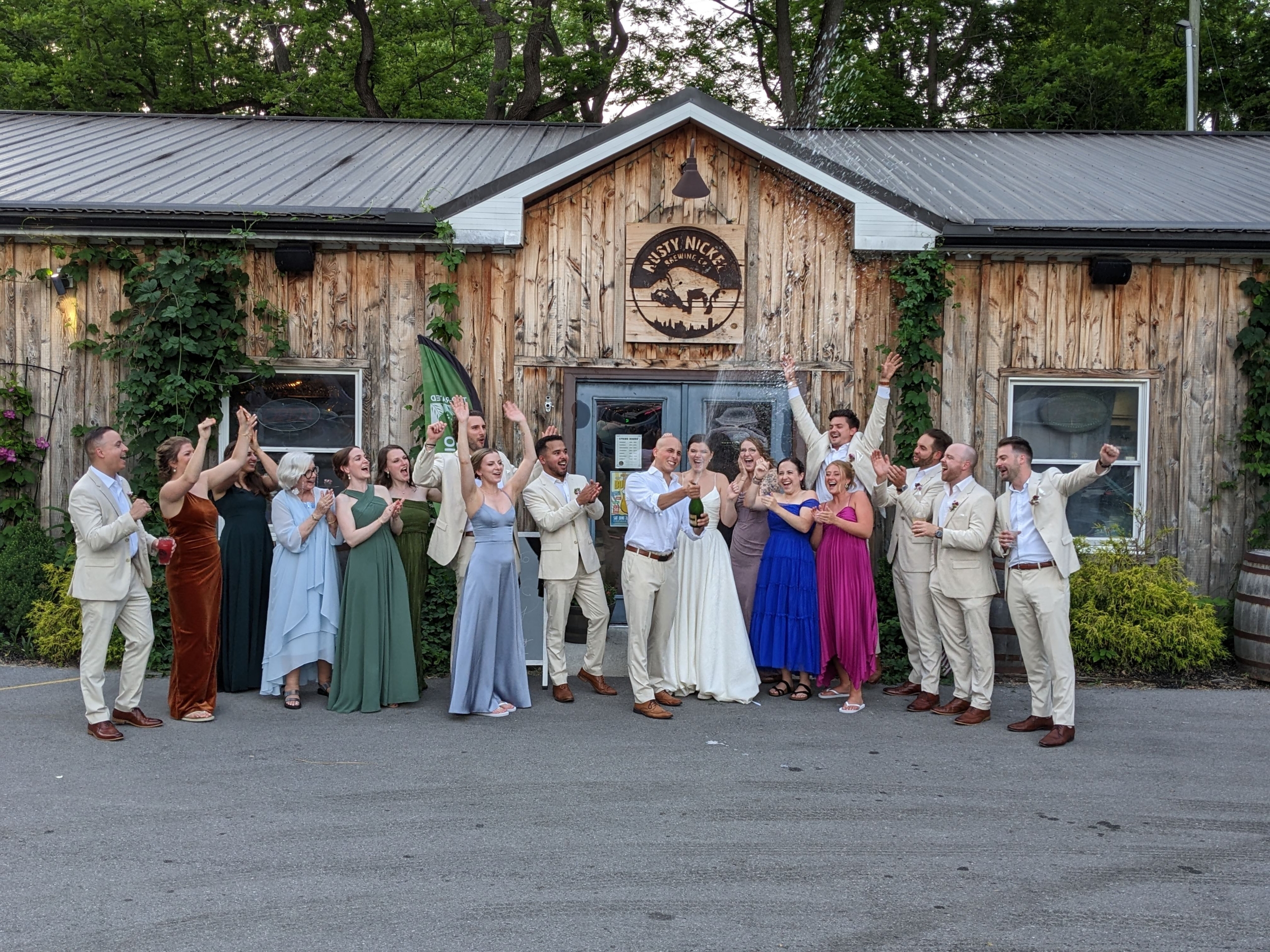 a wedding party cheers as a newlywed couple sprays champagne in celebration. The group is standing in front of Rusty Nickel Brewing.