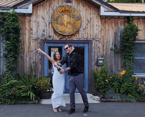 a bride jumps for joy as her groom sprays a bottle of champagne in celebration of their marriage. they are in front of the tasting room of Rusty Nickel Brewing.
