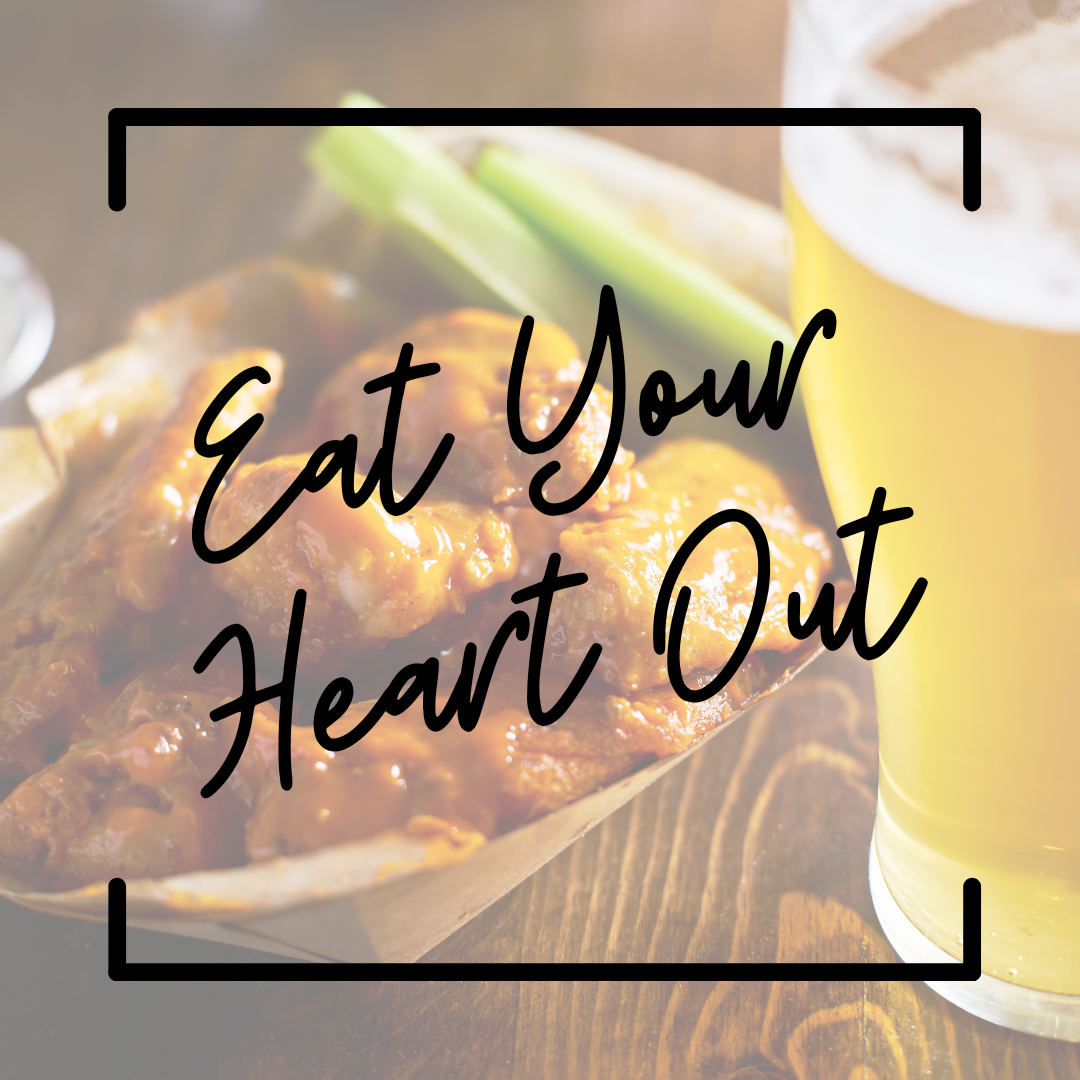 A wooden table top with a basket of chicken wings, celery, and a pint of beer. Text says "Eat Your Heart Out"