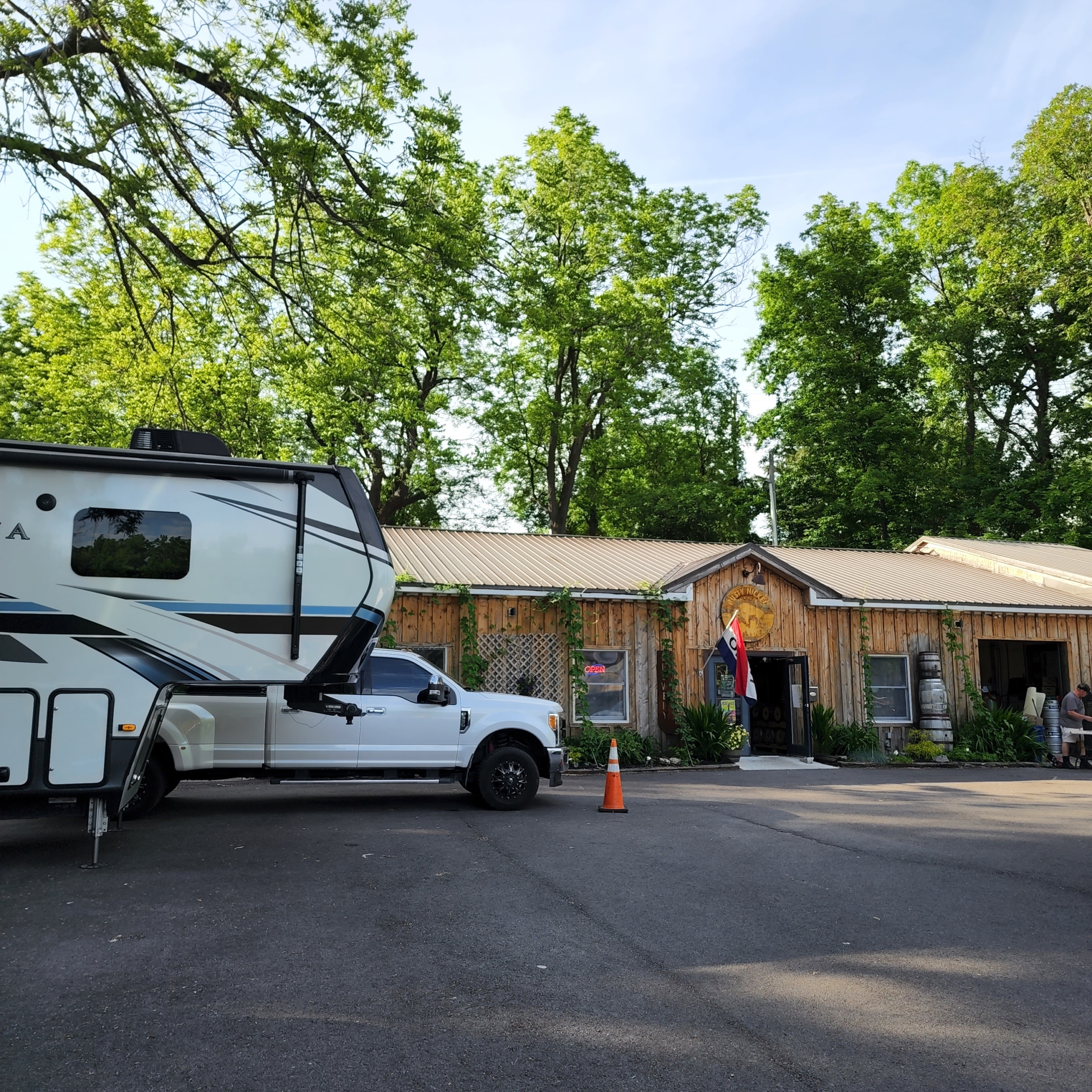 A travel trailer and pick-up truck are parked in designated Harvest Host spots in the parking lot in front of Rusty Nickel Brewing. They are shaded from the summer sun by large trees surrounding the grounds.