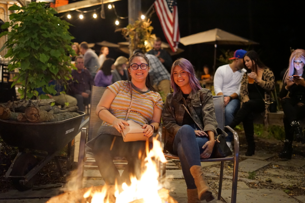 Two women sit by the firepit in the beer garden. The sky dark, but patio lights shine overhead.