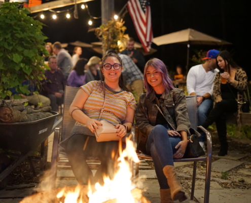 Two women sit by the firepit in the beer garden. The sky dark, but patio lights shine overhead.