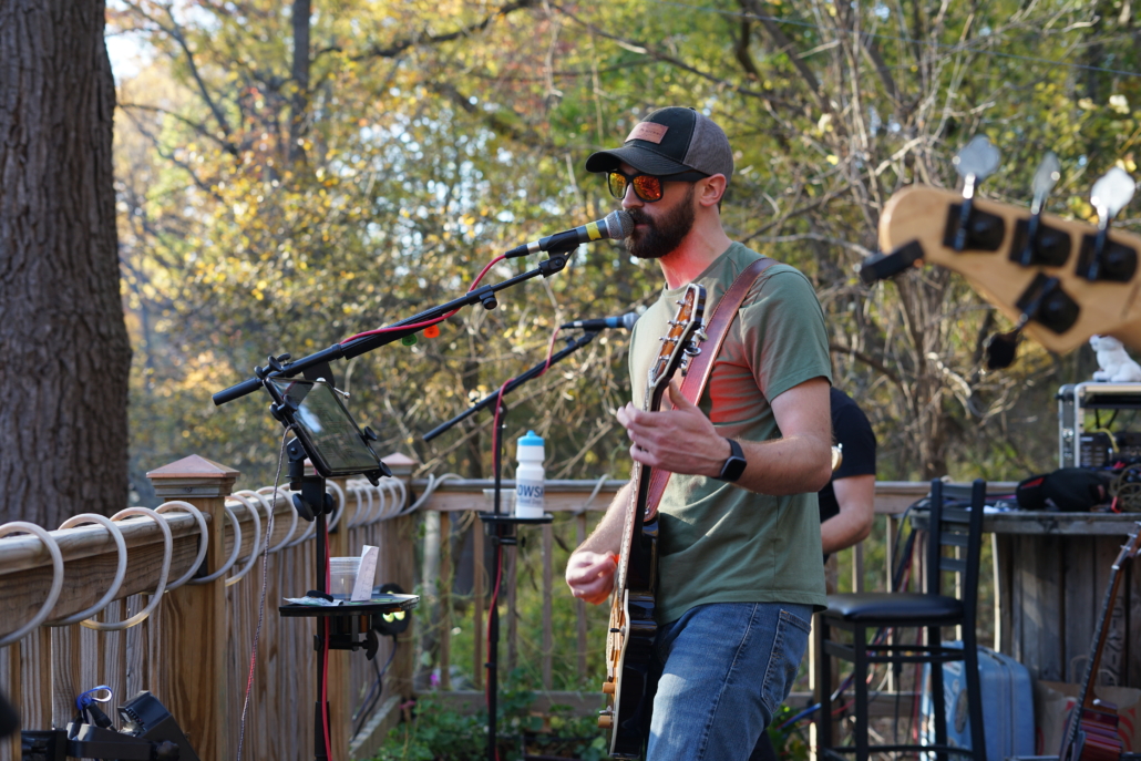 A musician performs on an outdoor stage on a sunny day. The musician is singing and playing guitar.