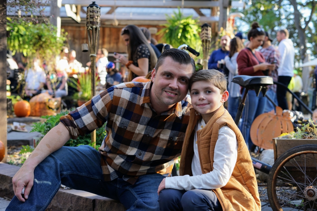 A father and young son pose for a photograph during a fall festival. The man is wearing a flannel and the boy is wearing a vest.