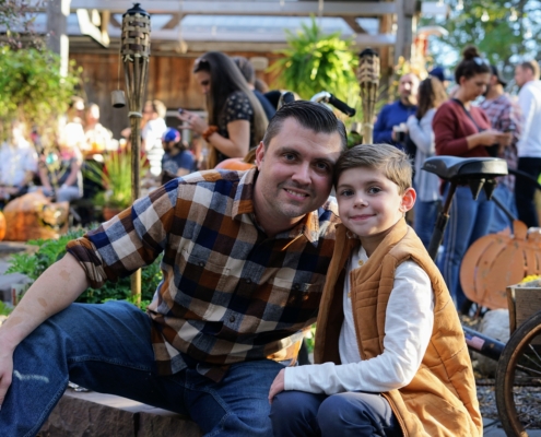 A father and young son pose for a photograph during a fall festival. The man is wearing a flannel and the boy is wearing a vest.