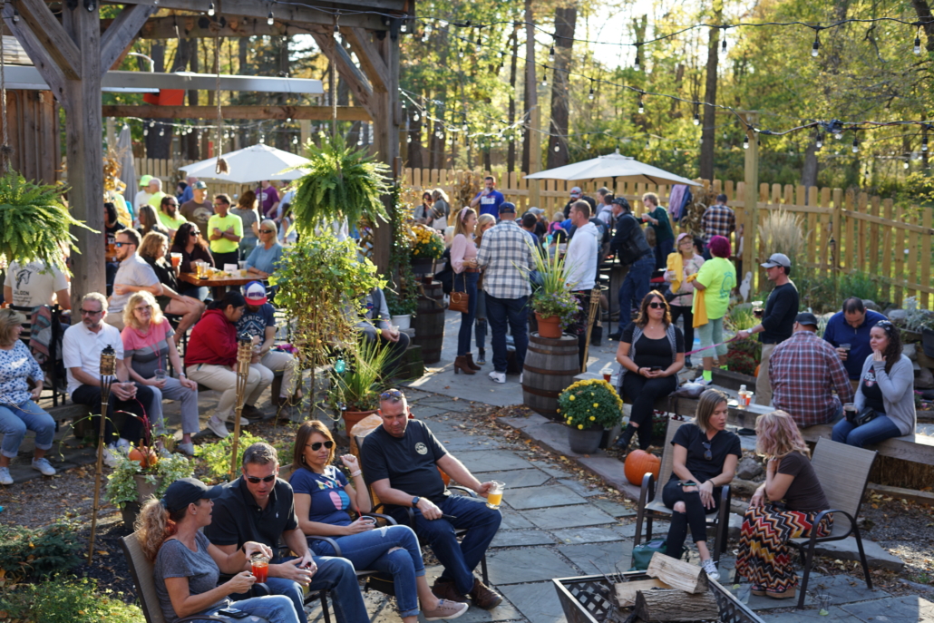 Guests enjoy the sunny patio and gardens during Harvest Fest at Rusty Nickel Brewing. Pumpkins, mums, and haybales are mixed in with garden beds and hanging plants. A firepit sits at the center of the stone pathway, with wood stacked in the middle. Guests are holding drinks and chatting.