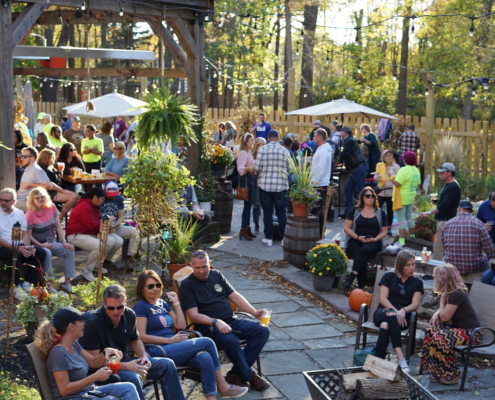 Guests enjoy the sunny patio and gardens during Harvest Fest at Rusty Nickel Brewing. Pumpkins, mums, and haybales are mixed in with garden beds and hanging plants. A firepit sits at the center of the stone pathway, with wood stacked in the middle. Guests are holding drinks and chatting.