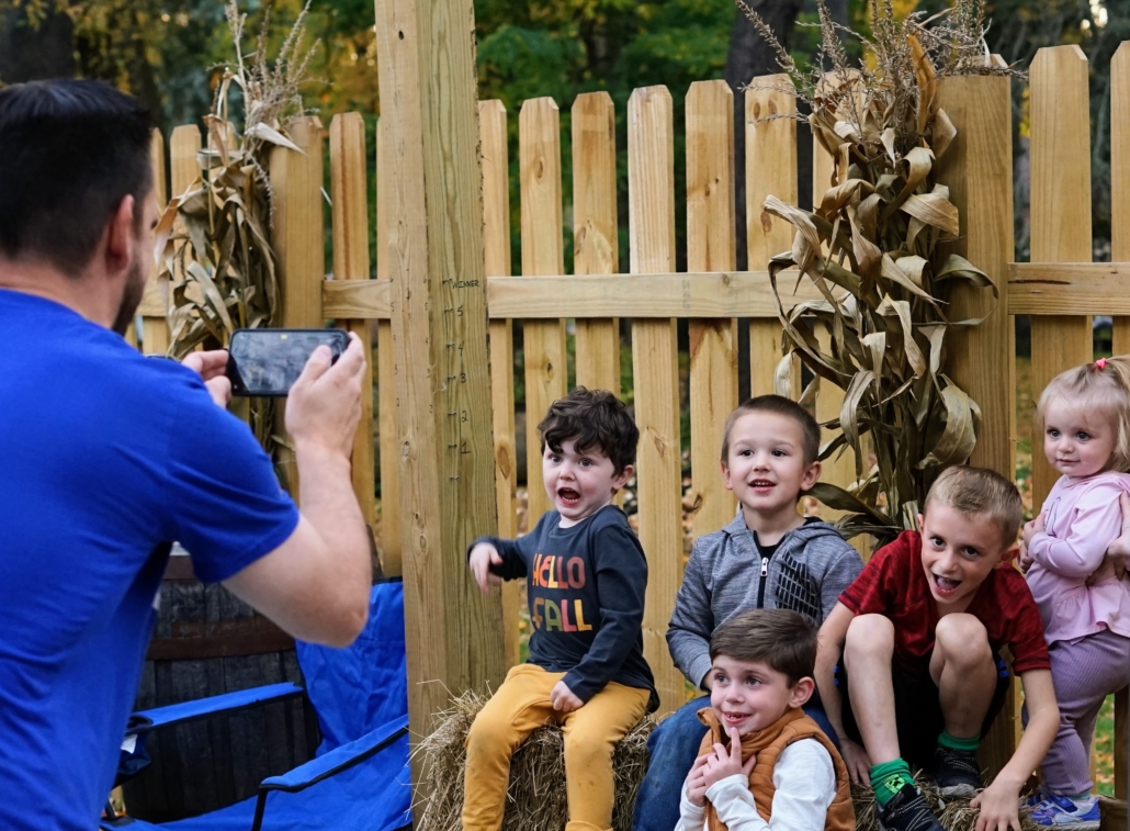 A man takes a photo of a group of kids sitting on haybales in front of a wooden fence. The area is decorated with cornstalks and pumpkins. The kids are laughing.