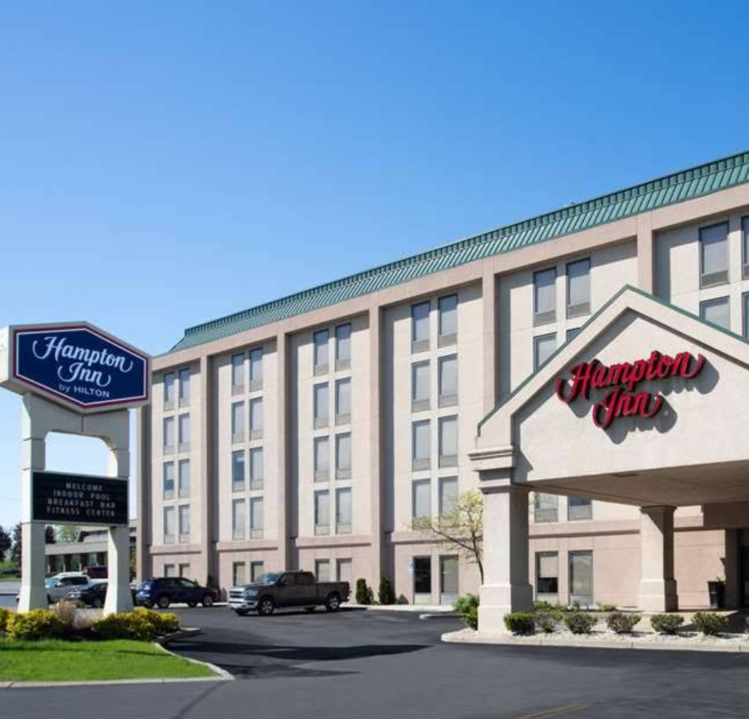 Exterior view of the Hampton Inn in West Seneca, NY. A large sign stands at the road. The building has a covered front entrance for vehicles.