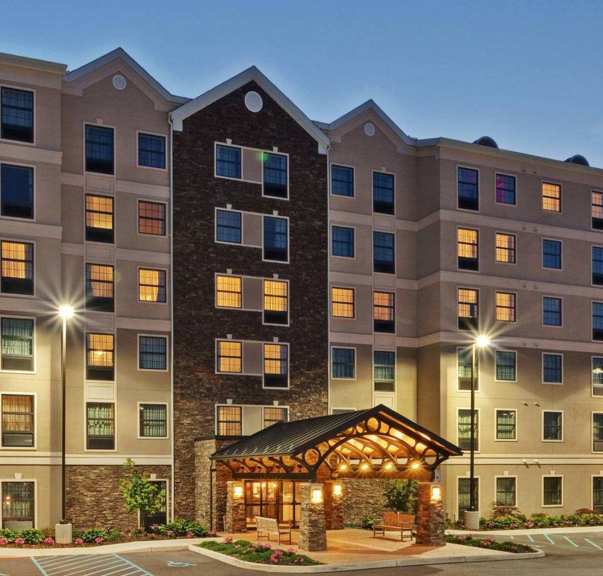 An exterior view of the Staybridge Suites in West Seneca, NY. The building is lit with exterior lighting. The walkway leading to the front entrance is covered with a large timber structure.
