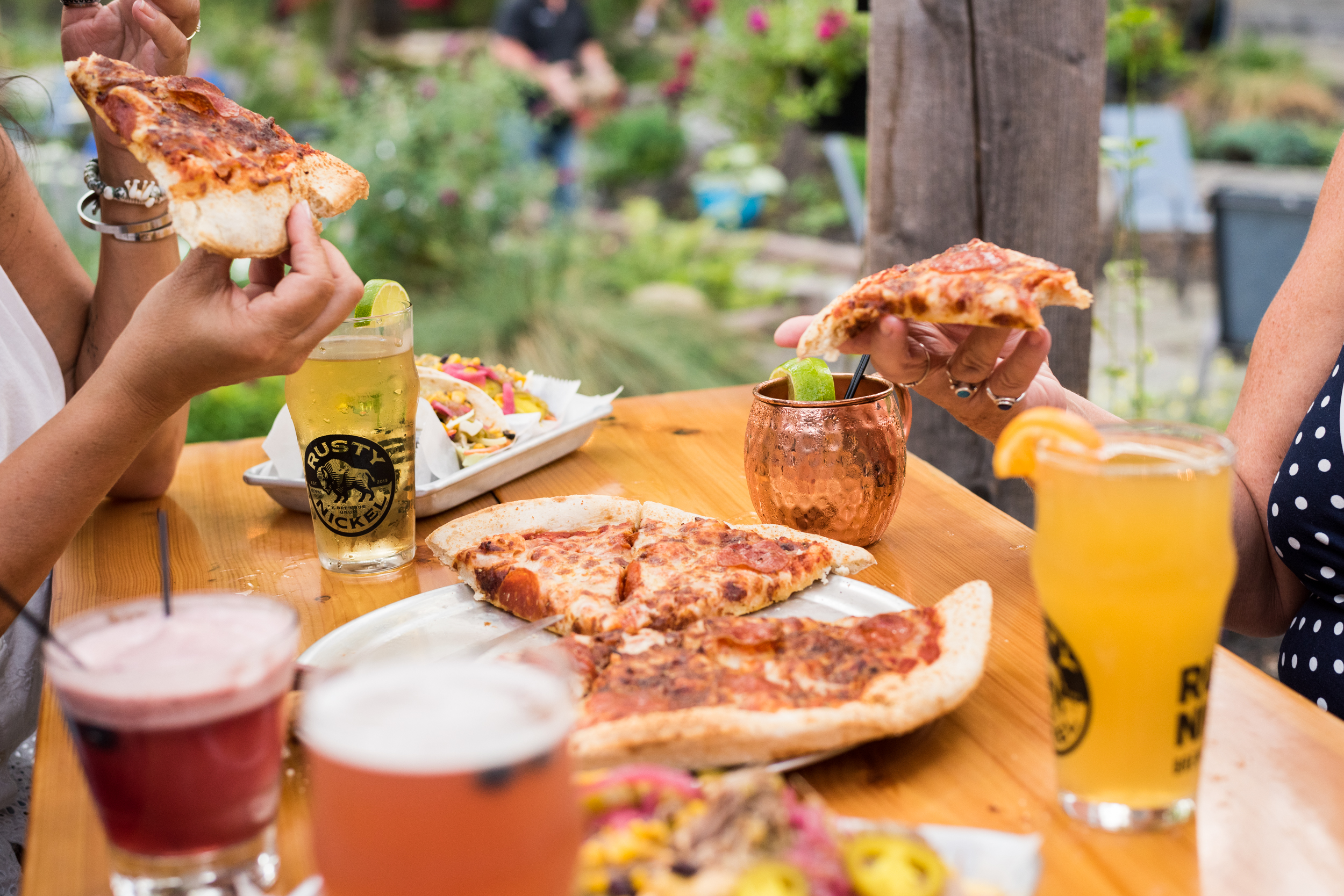 SOB-RustyNickel-080525-Selects-78 (1) a patio table top is filled with food and drinks being shared by patrons. A cheese and pepperoni pizza and nachos sit in the center, Beers and cocktails sit in front of each guest.